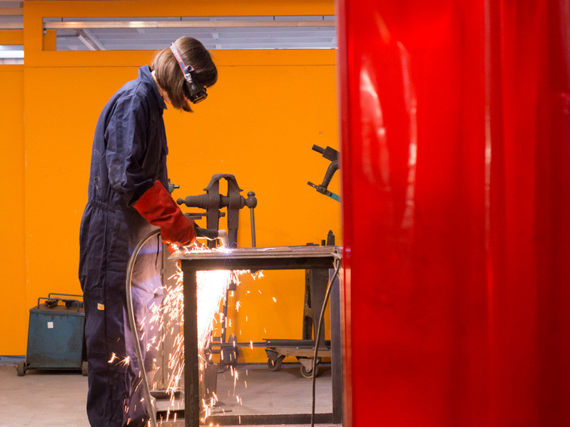 An artist in a blue boiler suit using a welding tool to join metal together
